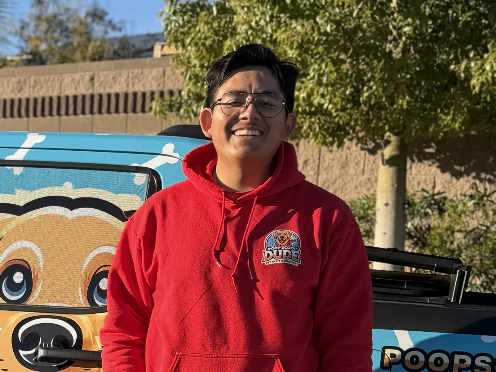 The owner of Poop Scoop Dude in the red company hoodie, standing in front of the wrapped truck in a Las Vegas neighborhood.